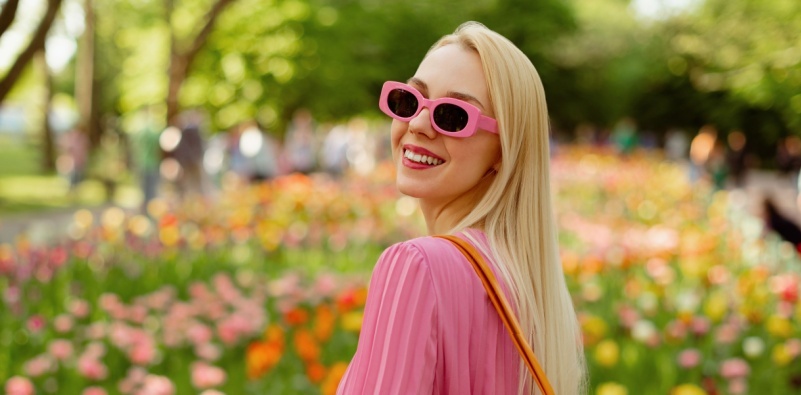- A woman smiling and wearing pink sunglasses walking in a park filled with flowers.