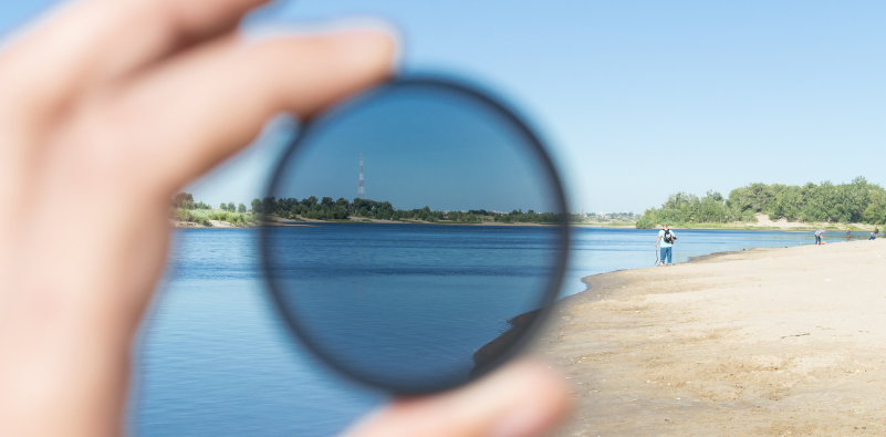 A person holding up a polarised lens to showcase reducing glare and improving clarity by the water