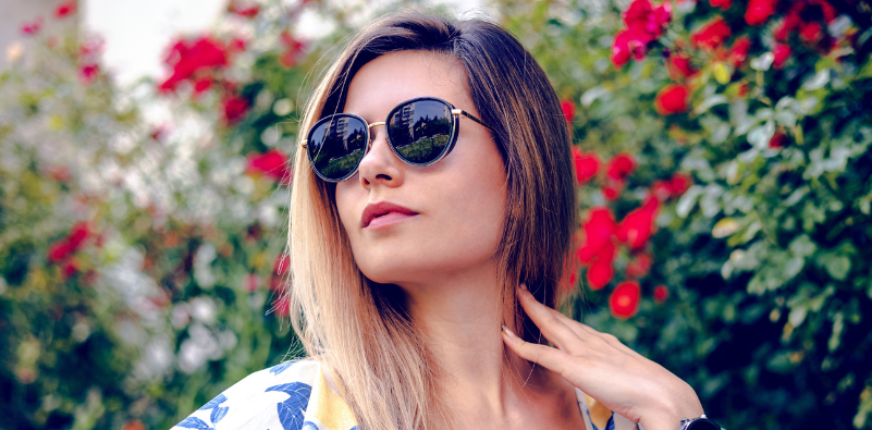 A young woman outside, surrounded by flowers, wearing a pair of round sunglasses